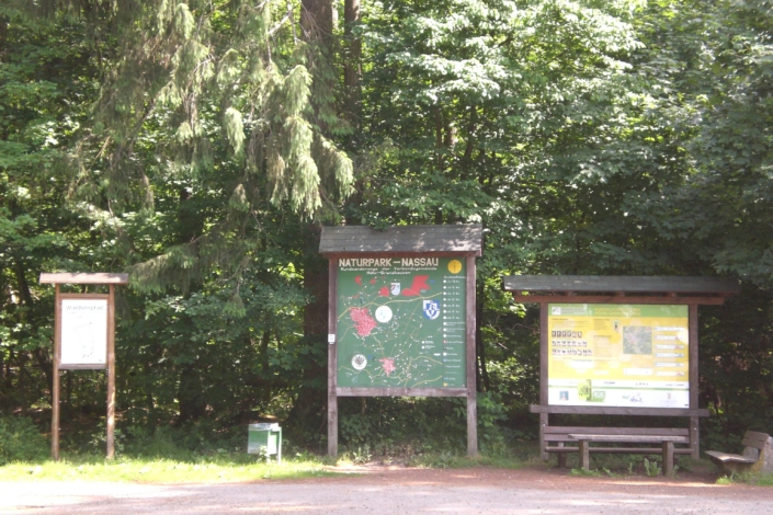 cimg1512 Drei große Informationstafeln und eine Bank am Eingang eines bewaldeten Naturparks, im Hintergrund Bäume.