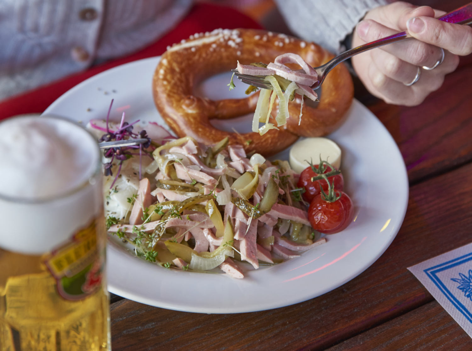 Eine Hand hält eine Gabel mit Salat über einem Teller mit Wurstsalat, Kirschtomaten und einer Brezel. Daneben steht ein Glas Bier.