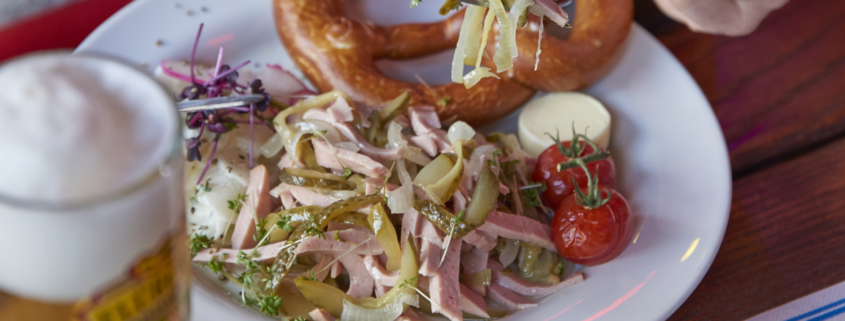 Eine Hand hält eine Gabel mit Salat über einem Teller mit Wurstsalat, Kirschtomaten und einer Brezel. Daneben steht ein Glas Bier.