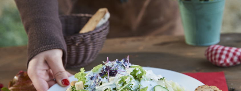 Eine Person serviert einen frischen Salat mit Gurken, Tomaten, Croutons und einer Scheibe Brot auf einem weißen Teller, der auf einem rustikalen Tisch steht.