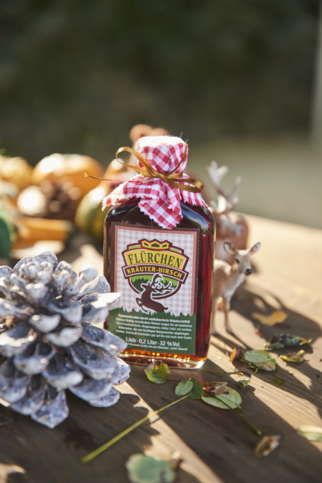 Eine Flasche Flürchen Kräuter-Hirsch-Likör mit kariertem Stoffverschluss steht neben einem Tannenzapfen und einem Spielzeughirsch auf einem Holztisch.