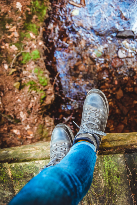 Eine Person in Bluejeans und Wanderstiefeln steht auf einer Holzkante über einem Bach, darunter sind Felsen und grünes Moos zu sehen.