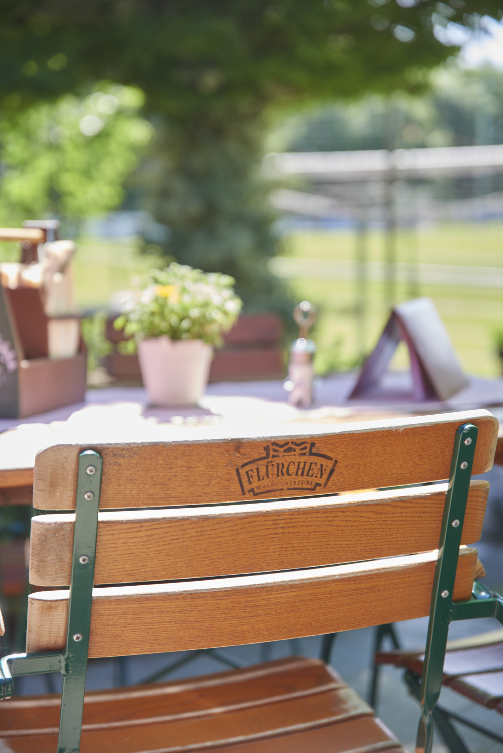 Holzstuhl an einem Cafétisch im Freien mit einer Topfpflanze, Speisekarten und Grünpflanzen im Hintergrund an einem sonnigen Tag.