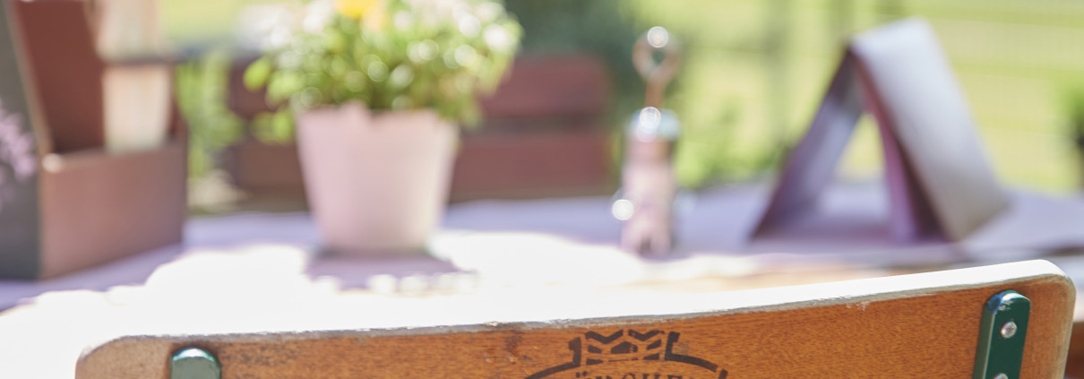 Holzstuhl an einem Cafétisch im Freien mit einer Topfpflanze, Speisekarten und Grünpflanzen im Hintergrund an einem sonnigen Tag.