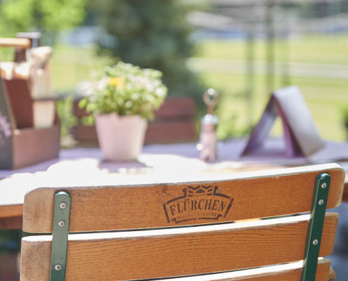 Holzstuhl an einem Cafétisch im Freien mit einer Topfpflanze, Speisekarten und Grünpflanzen im Hintergrund an einem sonnigen Tag.