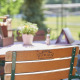 Holzstuhl an einem Cafétisch im Freien mit einer Topfpflanze, Speisekarten und Grünpflanzen im Hintergrund an einem sonnigen Tag.