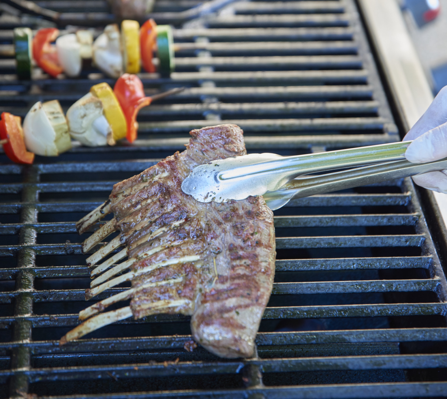 Eine Person grillt Lammkarree, Gemüsespieße und Würstchen auf einem Grill im Freien.