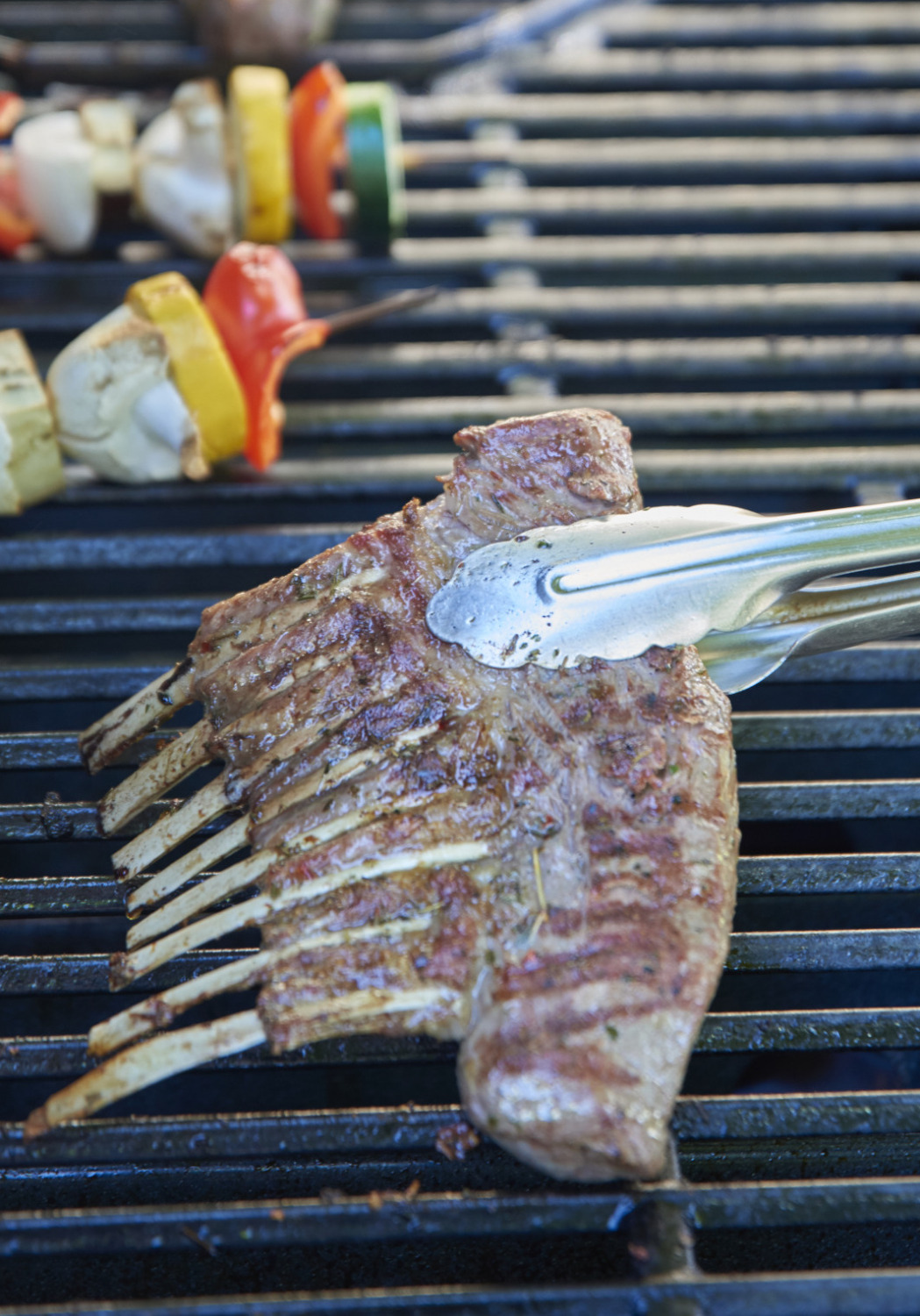 Eine Person grillt Lammkarree, Gemüsespieße und Würstchen auf einem Grill im Freien.