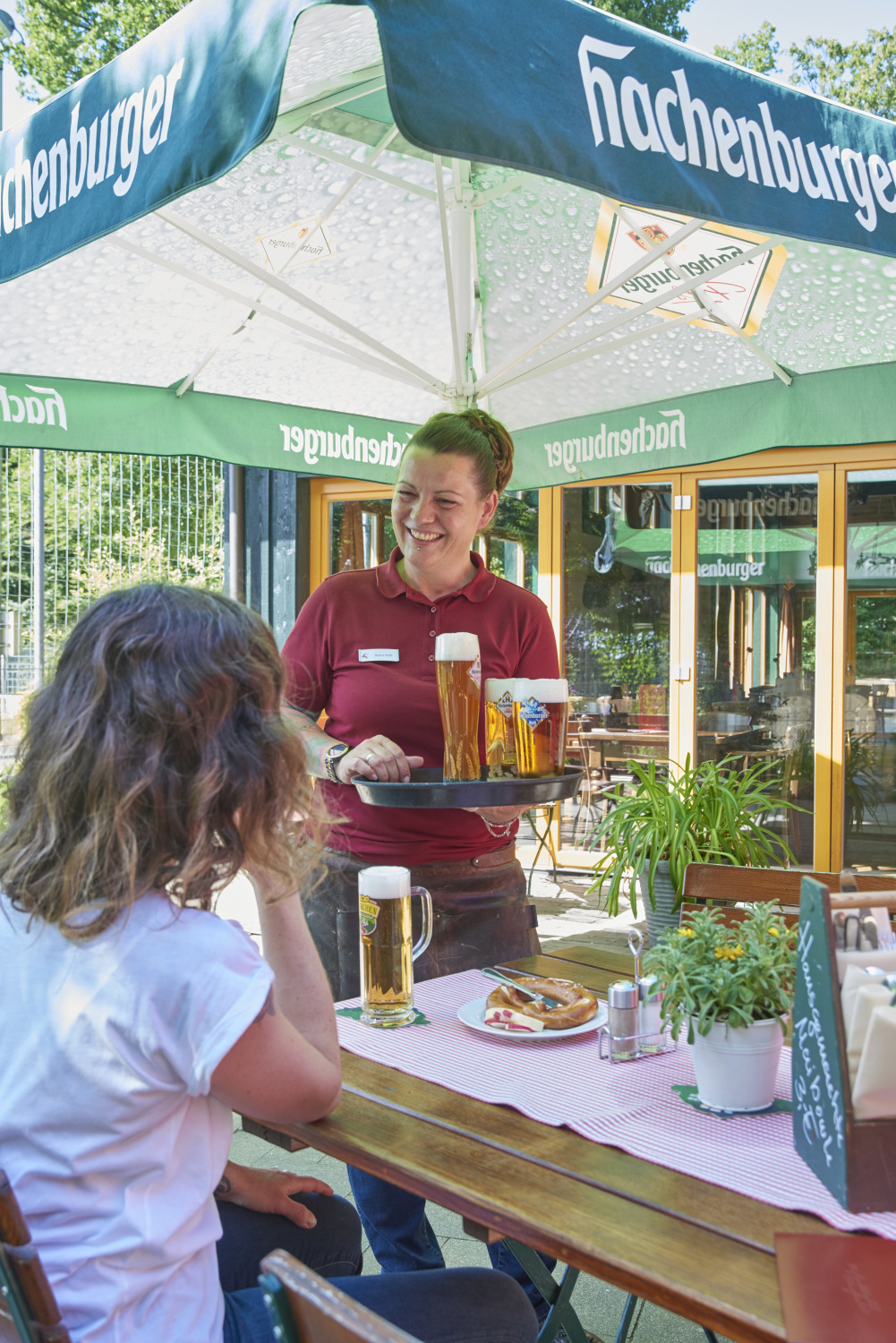 Ein lächelnder Kellner bringt einer Frau Bier, die mit Essen an einem Cafétisch im Freien unter einem großen Hachenburger-Sonnenschirm sitzt.