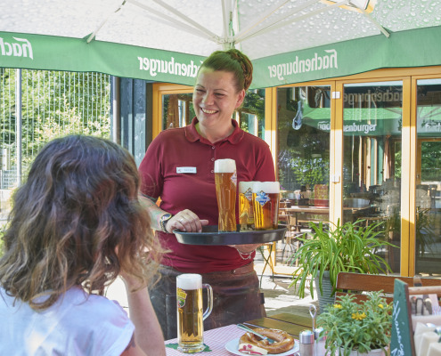 Ein lächelnder Kellner bringt einer Frau Bier, die mit Essen an einem Cafétisch im Freien unter einem großen Hachenburger-Sonnenschirm sitzt.