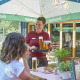 Ein lächelnder Kellner bringt einer Frau Bier, die mit Essen an einem Cafétisch im Freien unter einem großen Hachenburger-Sonnenschirm sitzt.