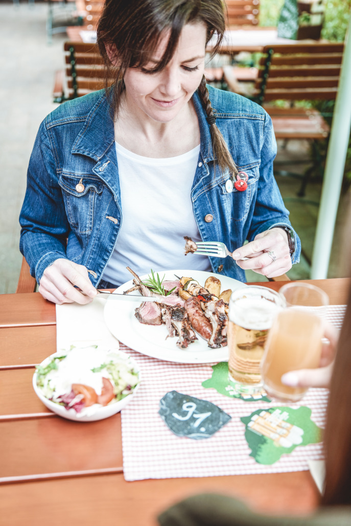 Eine Frau in einer Jeansjacke isst gegrilltes Fleisch mit Gemüse an einem Tisch im Freien, neben ihr stehen Bier und Salat.