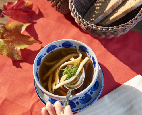 Eine Hand hält einen Löffel über einer Schüssel Nudelsuppe; Brotscheiben liegen in einem Korb und Herbstblätter liegen auf dem Tisch.