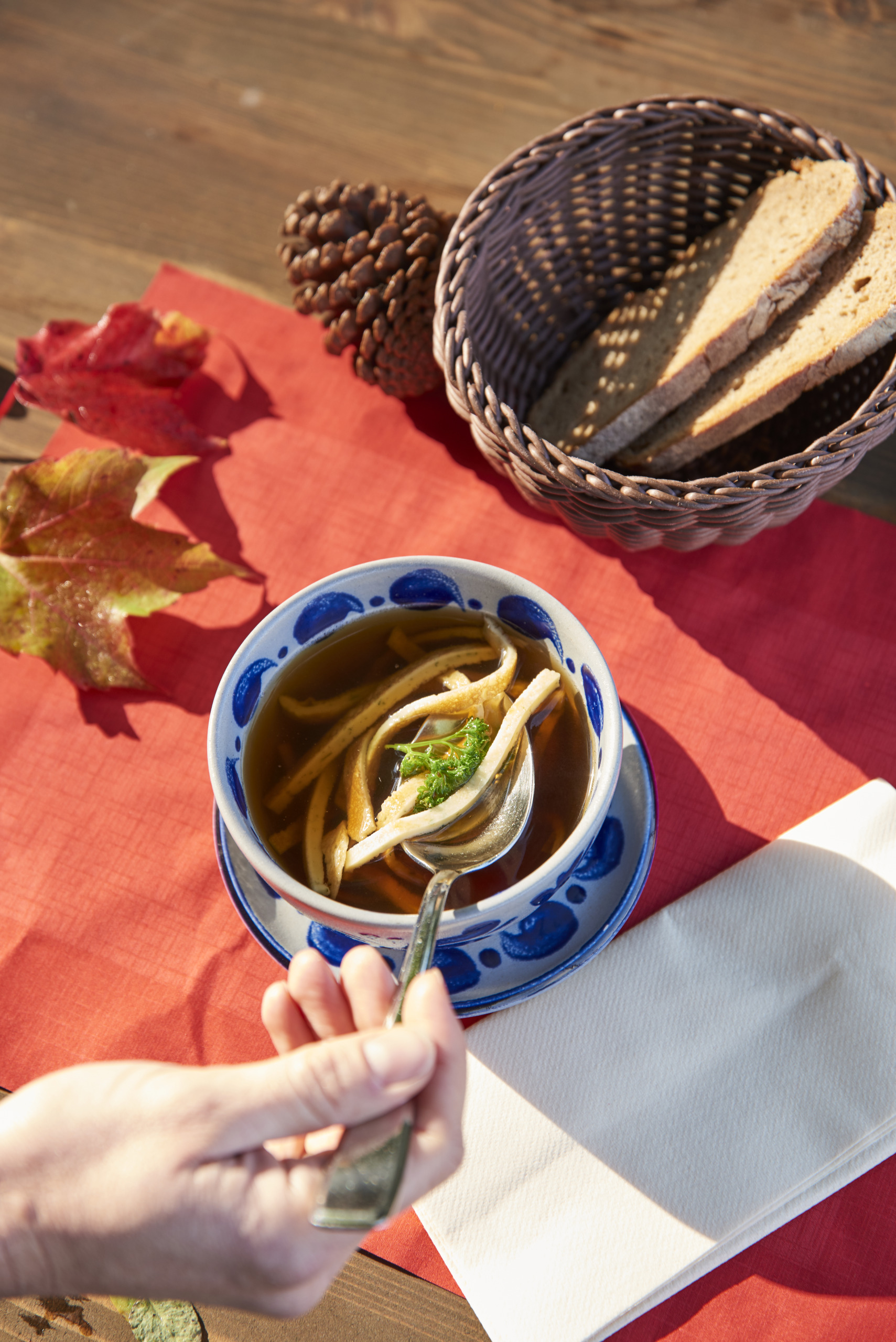 _BR18685 Eine Hand hält einen Löffel über einer Schüssel Nudelsuppe; Brotscheiben liegen in einem Korb und Herbstblätter liegen auf dem Tisch.