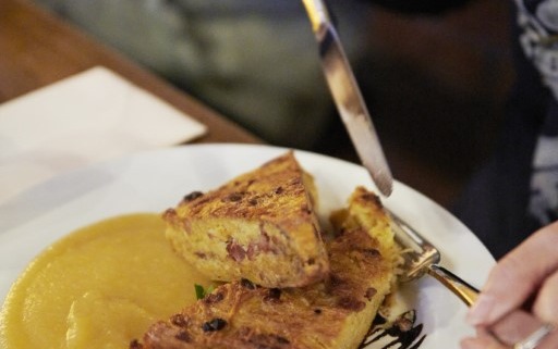 Eine Person schneidet eine Quiche in zwei Stücke, die mit einer Beilage Soße und einer Tomatengarnitur auf einem weißen Teller serviert wird.