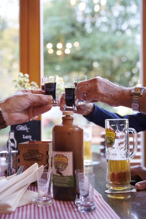 Drei Personen heben kleine Gläser mit dunklem Schnaps zum Anstoßen über einen Tisch mit Bier, einer Flasche und einer gestreiften Tischdecke.