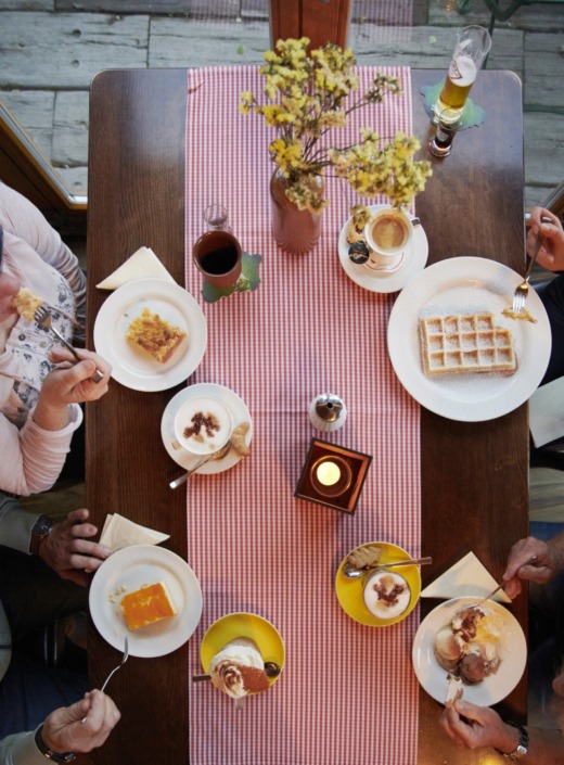 Ein für vier Personen gedeckter Tisch mit Desserts, Kaffee und Getränken auf einem rotkarierten Tischläufer. Man sieht Menschen, die Kuchen, Waffeln und Parfaits essen. In der Mitte stehen eine Kerze und eine Vase mit gelben Blumen.