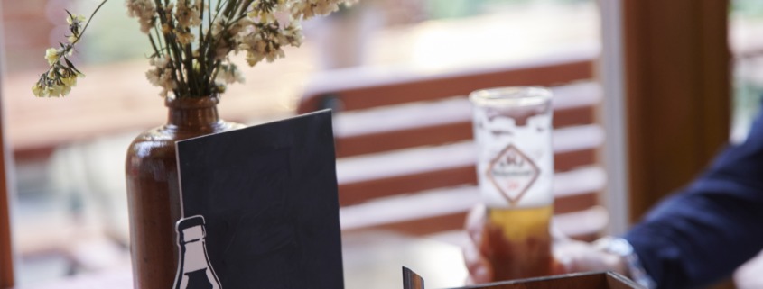 Ein mit Gewürzen, Servietten und einer Vase mit Blumen gedeckter Tisch in einem sonnendurchfluteten Café mit durch das Fenster sichtbaren Sitzgelegenheiten im Freien.