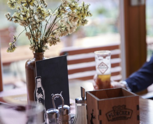 Ein Restauranttisch mit einer Blumenvase, Gewürzen, Servietten und einem Getränk; im Hintergrund sind Sitzgelegenheiten im Freien zu sehen.