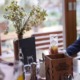 Ein Restauranttisch mit einer Blumenvase, Gewürzen, Servietten und einem Getränk; im Hintergrund sind Sitzgelegenheiten im Freien zu sehen.