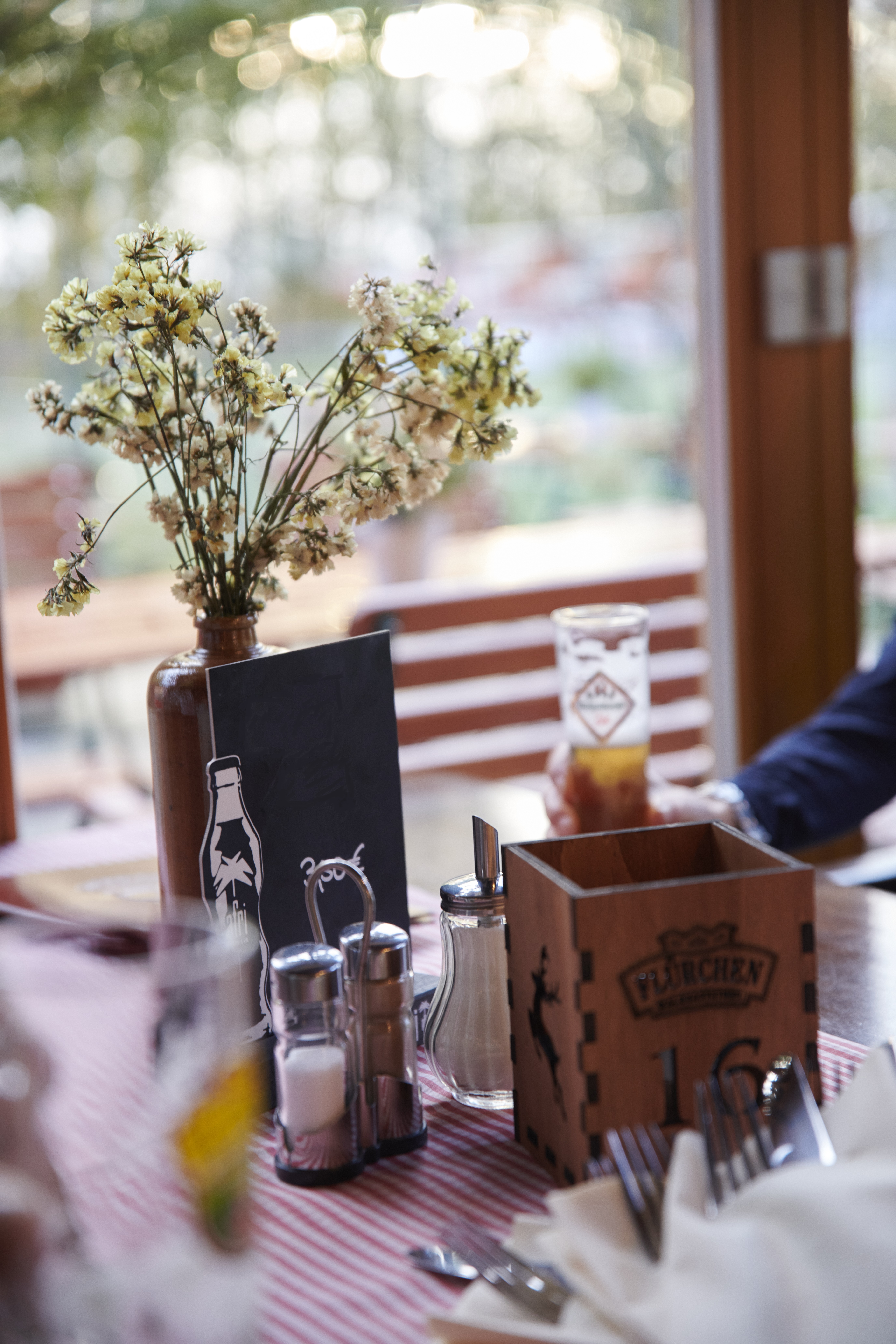 FL Tischdeko Tafel Hoch Ein Restauranttisch mit einer Blumenvase, Gewürzen, Servietten und einem Getränk; im Hintergrund sind Sitzgelegenheiten im Freien zu sehen.