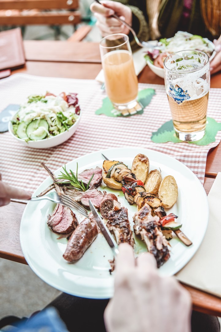 HotelHeinzMai23 899 Ein Teller mit gegrilltem Fleisch, Wurst, Gemüse und Kartoffeln auf einem Tisch mit Salat, Bier und Saft; Leute essen im Freien.