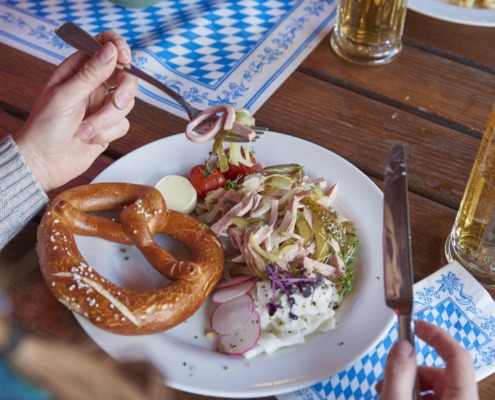 Eine Person hält das Besteck über einen Teller mit einer Brezel, Salat, Radieschen und Butter; auf dem Holztisch stehen ein Bier und eine Pflanze.