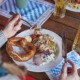 Eine Person hält das Besteck über einen Teller mit einer Brezel, Salat, Radieschen und Butter; auf dem Holztisch stehen ein Bier und eine Pflanze.