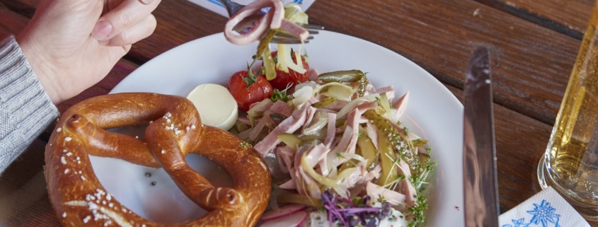Eine Person hält das Besteck über einen Teller mit einer Brezel, Salat, Radieschen und Butter; auf dem Holztisch stehen ein Bier und eine Pflanze.