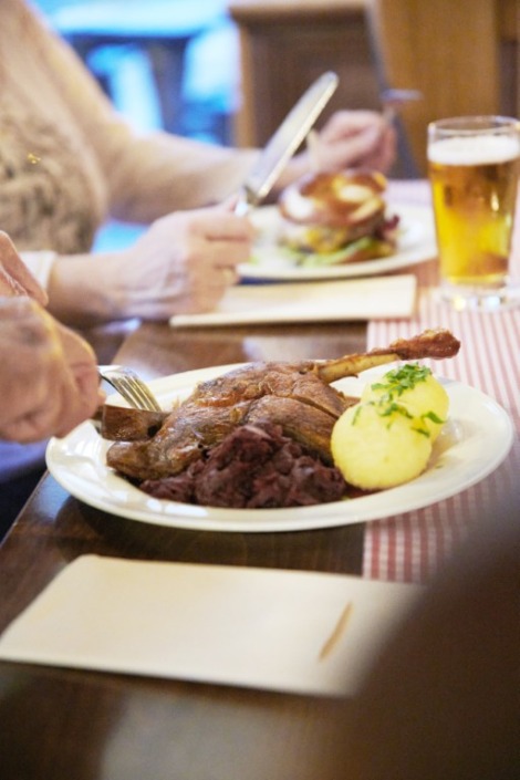 Eine Person schneidet einen Braten mit Kartoffeln und Rotkohl an, während im Hintergrund ein weiterer Teller und ein Glas Bier stehen.
