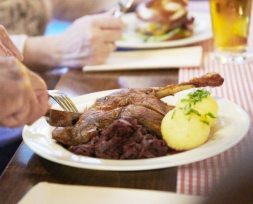 Eine Person schneidet einen Braten mit Kartoffeln und Rotkohl an, während im Hintergrund ein weiterer Teller und ein Glas Bier stehen.