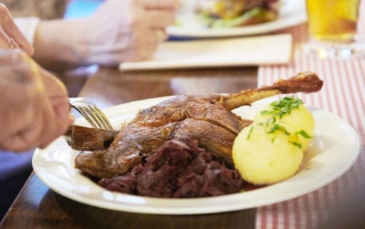 Eine Person schneidet einen Braten mit Kartoffeln und Rotkohl an, während im Hintergrund ein weiterer Teller und ein Glas Bier stehen.