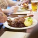 Eine Person schneidet einen Braten mit Kartoffeln und Rotkohl an, während im Hintergrund ein weiterer Teller und ein Glas Bier stehen.