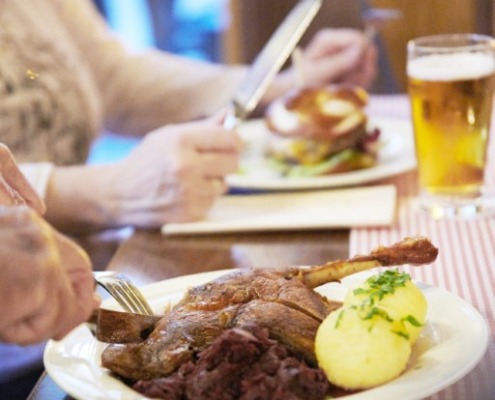 Eine Person schneidet einen Braten mit Kartoffeln und Rotkohl an, während im Hintergrund ein weiterer Teller und ein Glas Bier stehen.