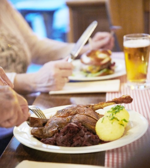 Eine Person schneidet einen Braten mit Kartoffeln und Rotkohl an, während im Hintergrund ein weiterer Teller und ein Glas Bier stehen.