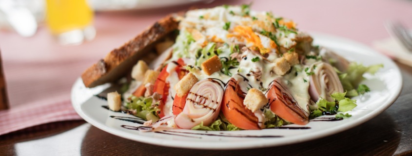 Ein Salatteller mit gerolltem Fleisch, Tomaten, Croutons, geriebenem Käse, Dressing und einer Scheibe getoastetem Brot als Beilage.